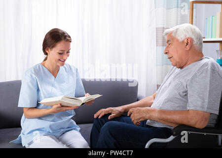 Nurse Reading Book For Disabled Senior Man Sitting On Wheelchair Stock Photo