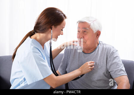 Smiling Young Nurse Examining Senior Man With Stethoscope At Home Stock Photo