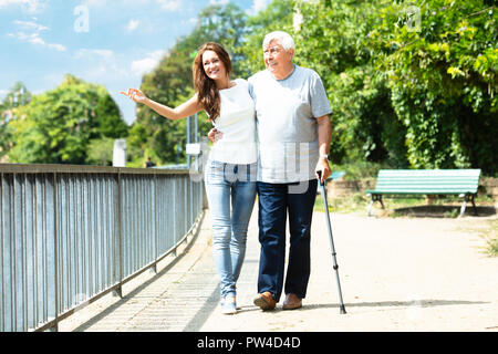 Young Woman Assisting Her Happy Father While Walking Near Railing In Park Stock Photo