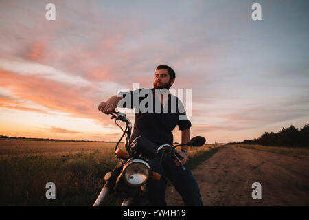 Thoughtful young man sitting on motorcycle at farm against sky during sunset Stock Photo
