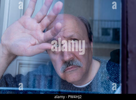 Old senior man looking throughout the window feeling confused and depressed alone at home in Aging Retirement Widower Dementia and Alzheimer concept. Stock Photo