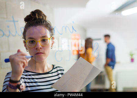businesswoman writing on window while colleagues discussing in background seen through glass Stock Photo
