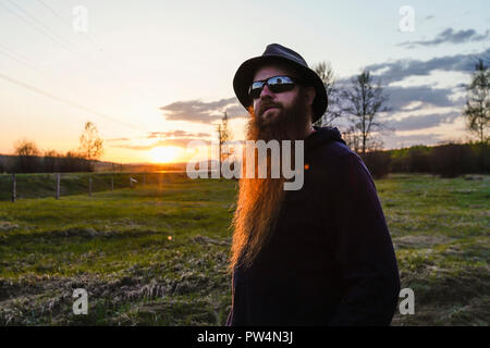 Bearded man wearing hat and sunglasses looking away while standing on field against sky during sunset Stock Photo
