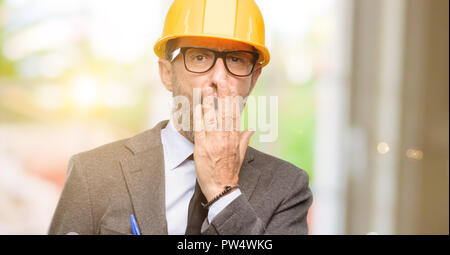 Senior architect or engineer covers mouth in shock, looks shy, expressing silence and mistake concepts, scared Stock Photo