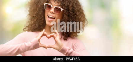 African american woman wearing pink sunglasses smiling in love showing heart symbol and shape with hands. Romantic concept. Stock Photo