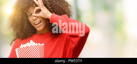 African american woman eating popcorn with happy face smiling doing ok sign with hand on eye looking through fingers Stock Photo