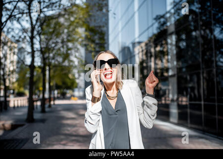 Excited business woman in white suit talking with smartphone at the financial district with modern buildings on the background Stock Photo