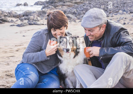 happy couple smiling and drink tea or coffee staying sit down closer with dog border collie in the middle. love and family alternative lifestyle conce Stock Photo
