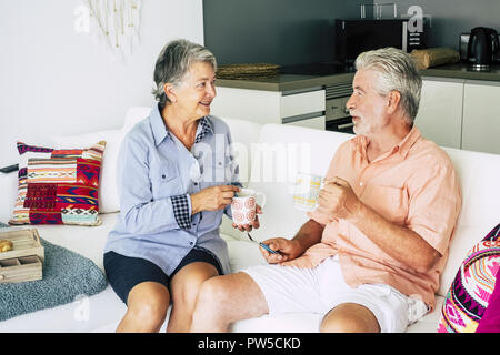 cheerful and relaxed couple of adult senior caucasian people at home sit down on a white sofa and speak together drinking a tea break. retired life co Stock Photo