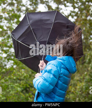 Kidderminster, UK. 12th October, 2018. UK weather: it's very wet and windy! Milder temperatures may be tempting us outdoors, but visitors to a local park in Kidderminster have to brave heavy spells of rain and increasingly strong, gusting winds this morning. An isolated young lady, rear view, is struggling in the wind: her long hair being blown about and her big, black brolly blown completely inside-out by a sudden gust of wind. Credit: Lee Hudson/Alamy Live News Stock Photo