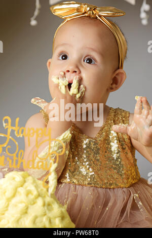 small sweet baby girl in a golden dress with a bow on her head trying a jazzy jelly cake from a cream. studio shot of a birthday on a gray background surrounded by balls Stock Photo