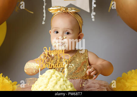 small sweet baby girl in a golden dress with a bow on her head trying a jazzy jelly cake from a cream. studio shot of a birthday on a gray background surrounded by balls Stock Photo