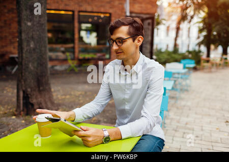 Young businessman enjoys coffee in outdoor cafe using tablet. Handsome guy drinking tea on city street holding tablet Stock Photo