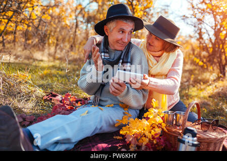 Senior couple using phone while having tea in autumn forest. Happy man and woman talking and pointing at screen on picnic Stock Photo