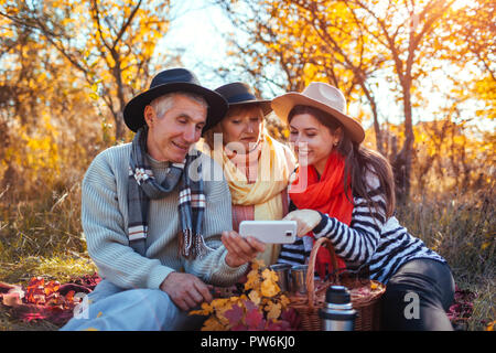 Senior parents using smartphone in autumn forest with their daughter. Family values. People having picnic together Stock Photo