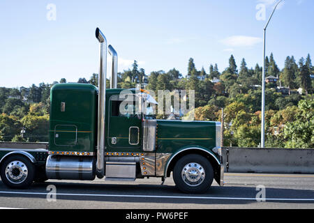 Profile of green classic American pro stylish bonnet big rig semi truck with high vertical exhaust pipes transporting commercial goods and driving on  Stock Photo