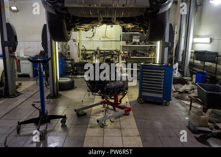 A sports car raised on a lift for repair and under it a detached engine suspended on a blue crane and a gear box on a lifting table in a vehicle repai Stock Photo