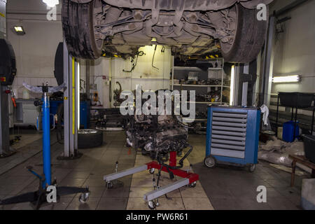 A sports car raised on a lift for repair and under it a detached engine suspended on a blue crane and a gear box on a lifting table in a vehicle repai Stock Photo