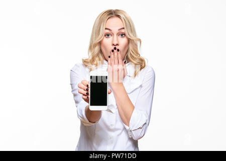 Portrait of shocked pretty blond business woman with hand on her mouth showing mobile phone blank screen isolated over white background. Shock, disbel Stock Photo