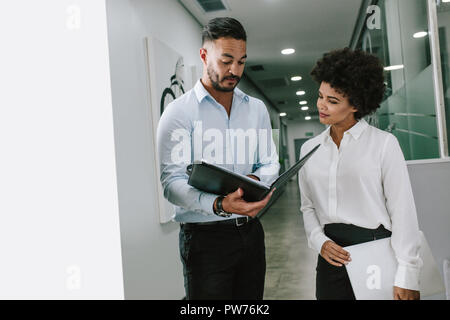 Businessman showing a file to female colleague on office corridor. Two business colleagues standing in office corridor and looking at a document. Stock Photo