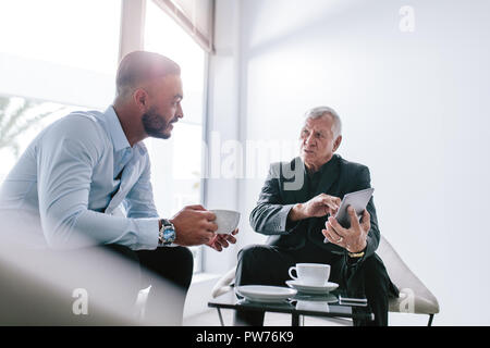Two businessman sitting in office lobby discussing work over a cup of coffee. Business colleagues talking during work break in office lobby. Stock Photo