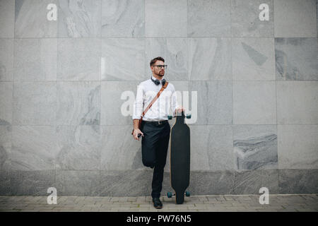 Formally dressed man standing in street with a longboard. Businessman standing against a wall outdoors holding a long skateboard. Stock Photo