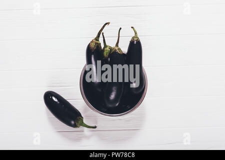 Raw purple eggplants on white wooden table in metal bowl, top view. Flat lay composition of group harvest aubergine vegetables. Stock Photo