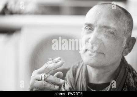 A 50y man is smoking a cigarette. Black and white. The fan coil of an air-conditioner is on the background. Stock Photo