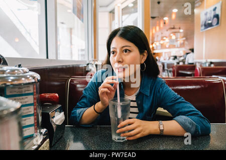 elegant lady sitting in the diner and drinking water with straw waiting for someone to come. young woman sitting in indoor restaurant during spring tr Stock Photo