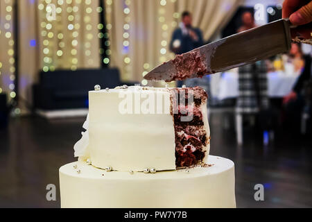 Cutting the first tier of a white mastic wedding cake in a banquet hall background. Wedding cake cutting sweet moment. Wedding traditions. cake etique Stock Photo