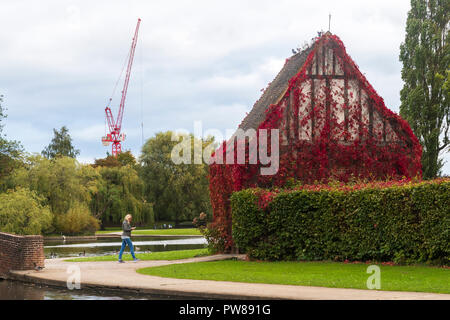 Autumn view of young woman walking & looking at phone by lake & lych-gate - beautiful, peaceful, municipal Rowntree Memorial Park, York, England, UK. Stock Photo