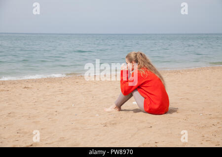 blonde in a Red Sweater is drinking hot coffee or tea on the sandy beach Stock Photo