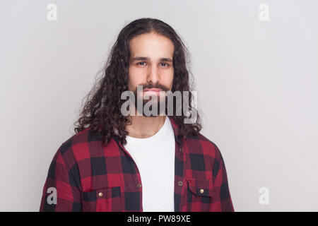Portrait of sad handsome man with beard and black long curly hair in casual style, checkered red shirt standing and looking at camera with unhappy fac