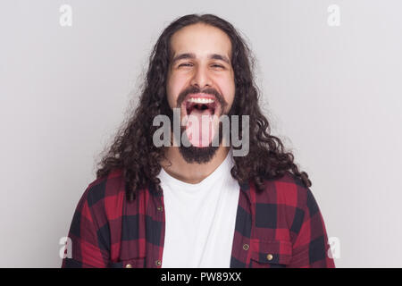 Crazy handsome man with beard and black long curly hair in casual style, checkered red shirt standing, open big mouth, tongue out and looking at camer Stock Photo