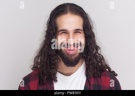 Funny crazy brunette man with beard and black long curly hair in casual style, checkered red shirt standing looking at camera and licking his nose. in Stock Photo