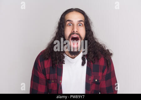 Portrait of amazed handsome man with beard and black long curly hair in casual checkered red shirt looking at camera with open mouth and big eyes. ind Stock Photo