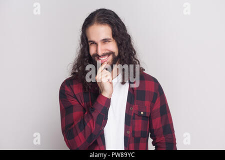 Portrait of playful handsome man with beard and black long curly hair in casual style, checkered red shirt standing looking at camera with cunning fac Stock Photo
