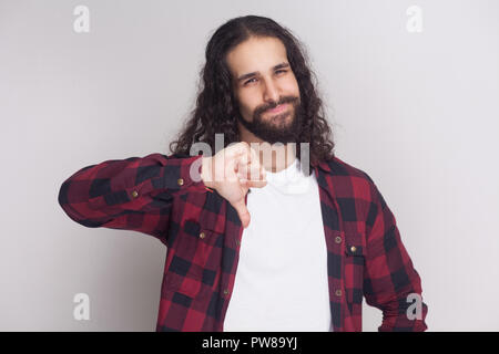 Portrait of unhappy handsome man with beard and black long curly hair in casual checkered red shirt standing looking at camera, showing dislike sign.  Stock Photo