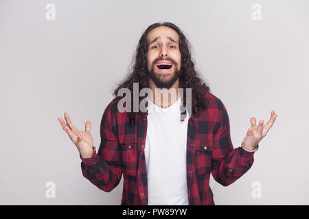 what i must to do? sad unhappy man with beard and black long curly hair in checkered red shirt standing with raised arms, looking at camera and asking