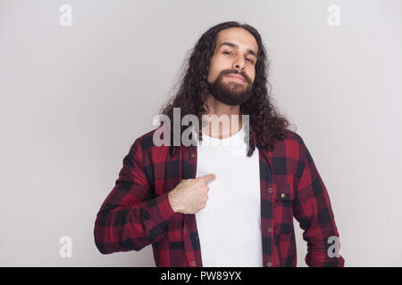 This is me. Portrait of proud handsome man with beard and black long curly hair in checkered red shirt standing, looking at camera and pointing himsel Stock Photo