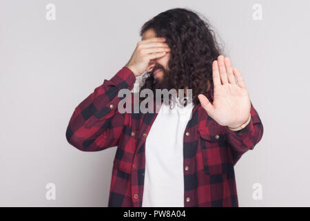 I dont want to see this. Portrait of handsome man with beard and black long curly hair in checkered red shirt standing covering eyes and stop gesture.