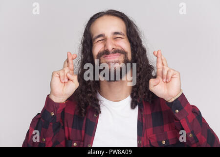 Portrait of hopeful handsome man with beard and black long curly hair in casual style, checkered red shirt standing closed eyes with crossed fingers.  Stock Photo