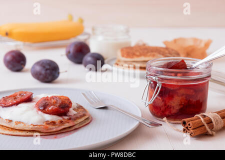 White wooden table with closeup image of dessert saucer with crumpets, pancakes, sour cream and fresh plums. The glass with cooked plums, fresh plums  Stock Photo