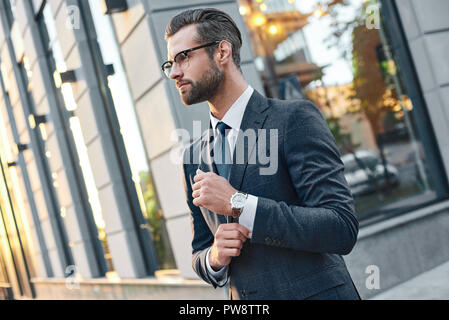 Close up profile portrait of a successful young bearded guy in suit and glasses. So stylish and nerdy. Outdoors on a sunny street, fixing his cuffs Stock Photo