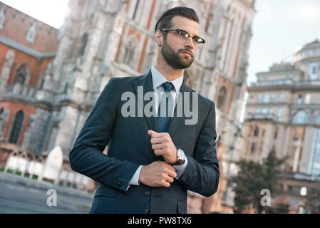 Close up profile portrait of a successful young bearded guy in suit and glasses. So stylish and nerdy. Outdoors on a sunny street, fixing his cuffs Stock Photo