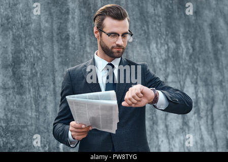 Young businessman in suit and glasses looks at the clock and reading business newspaper in his hands Stock Photo