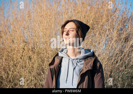Young woman enjoys the autumn sun. Female in parka outdoors on bright sunny afternoon