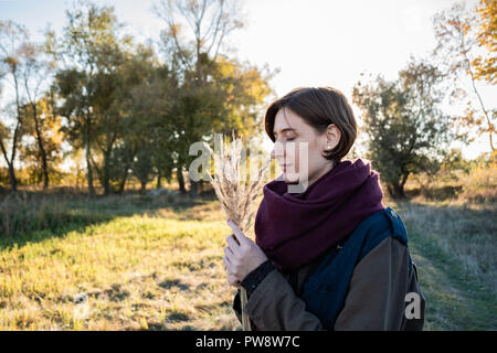 Young woman with a bunch of field grasses enjoying beautiful late summer day. Portrait of female with closed eyes in backlit autumn background Stock Photo