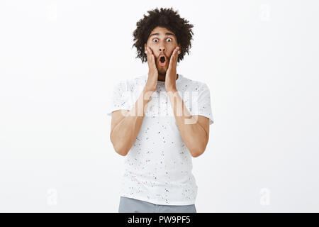 Man screaming from shock, being impressed favorite band comming in town. Portrait of amazed good-looking hispanic guy in casual t-shirt, folding lips with opened mouth, holding palms on cheeks Stock Photo