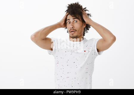 Guy realised he has son, being shocked and standing in stupor. Potrait of stunned confused and astonished funny dark-skinned male, touching hair and standing back with scared expression over grey wall Stock Photo